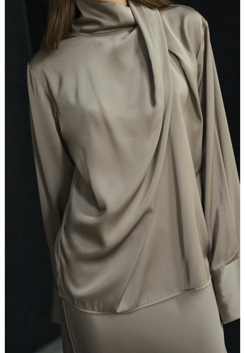 Femme portant un chemisier ample en satin taupe avec un col montant et des manches longues, visible des épaules aux hanches sur un fond sombre.