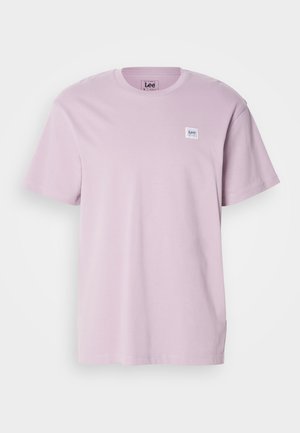 Camiseta de manga corta de color púrpura claro, hecha de algodón, con un pequeño parche logotipo rectangular blanco en la zona del pecho izquierdo. Textura suave.