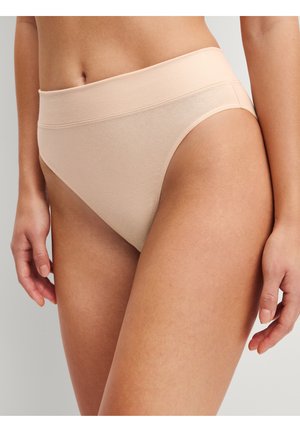 Femme portant des sous-vêtements en coton beige taille haute, montrant le torse et le haut des cuisses sur un fond uni.
