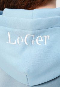 Ανοιχτό μπλε φούτερ με κουκούλα που φοράει άτομο με κόκκινα σγουρά μαλλιά, με λευκό κεντημένο κείμενο "LeGer" στην πίσω πλευρά της κουκούλας.