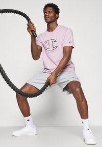 Helles lila Baumwoll-T-Shirt mit einem schwarzen Logo, graue Sportshorts, weiße Turnschuhe und ein schwarzes Battle Rope vor einem neutralen Hintergrund.