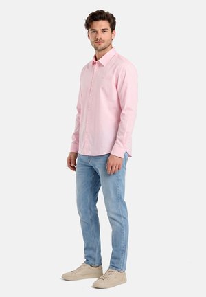 Hombre de pie vistiendo una camisa de manga larga con botones de color rosa claro, vaqueros azul claro y zapatillas beige sobre un fondo liso.