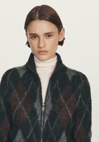 Cardigan con zip di colore verde scuro e marrone con un motivo a diamante. Tessuto morbido e testurizzato con colletto alto e design aderente.