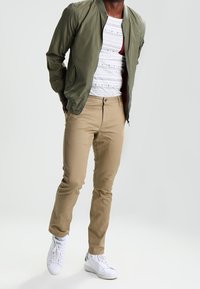Giubbotto bomber verde oliva sopra una maglietta bianca a motivi, pantaloni khaki e sneaker bianche con dettagli verdi.