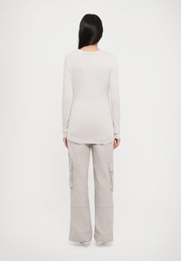 Femme aux longs cheveux bruns se tient de dos, portant un haut à manches longues beige clair, un pantalon cargo assorti et des talons beiges.