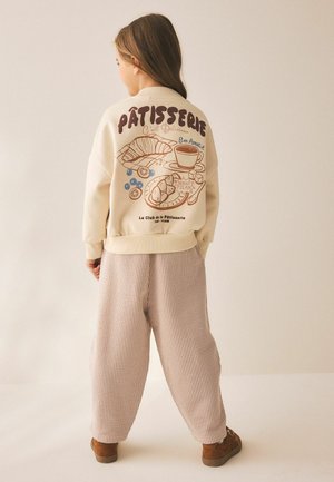Kind trägt cremefarbene Jacke mit Patisserie-Illustrationen und Text, lockere beige Hose und braune Schuhe, steht mit dem Rücken zum Betrachter auf einfarbigem Hintergrund.