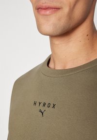 Olíva zöld pulóver kerek nyakkivágással, fekete hímzett "HYROX" felirattal és egy kis fekete Puma logóval a mellkasán. Simaságával tűnik ki.