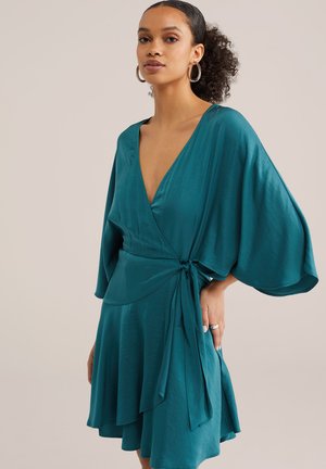 Mlada ženska z kodrastimi lasmi v modro-zelena obleka z ohlapnimi rokavi, vezana okoli pasu, stoji pred enobarvnim bež ozadjem.