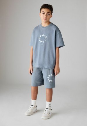 Teen boy stands wearing blue matching T-shirt and shorts with circular "SAINTS LONDON" logos, white socks, and white sneakers.