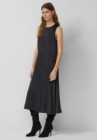 Zwarte mouwloze jurk met een gladde textuur, A-lijn silhouet en diagonale naaddetails, gecombineerd met zwarte laarzen met hoge hakken.