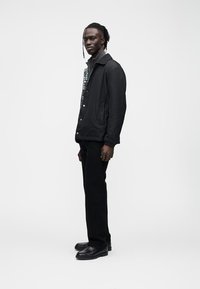 Casaco preto com um design estruturado, apresentando um detalhe estampado no lado esquerdo, combinado com calças pretas e sapatos de couro.