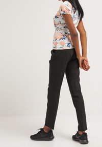 Kobieta stoi z dłońmi splecionymi za plecami, ubrana w kwiecisty t-shirt z krótkim rękawem, czarne spodnie i czarne sneakersy, na jednolitym tle.