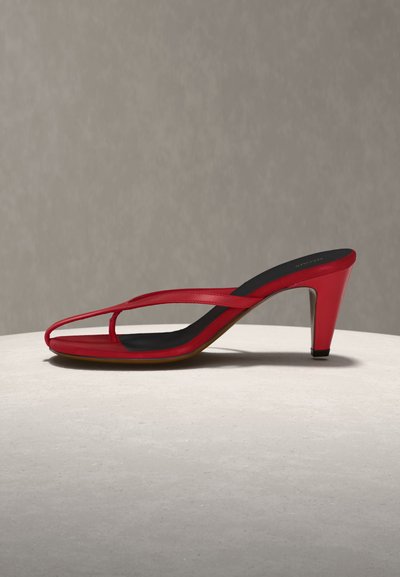 Κόκκινο σανδάλι με λεπτές λουρίδες, τετράγωνη μύτη και μαύρη σόλα, τοποθετημένο σε ανοιχτόχρωμη επιφάνεια με ουδέτερο φόντο.
