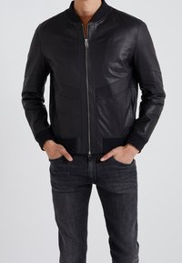 Czarna skórzana kurtka bomberka z przednim zamkiem błyskawicznym, ściągaczami przy mankietach i u dołu oraz pionowymi przeszyciami podkreślającymi strukturę. W połączeniu z ciemnymi jeansami.