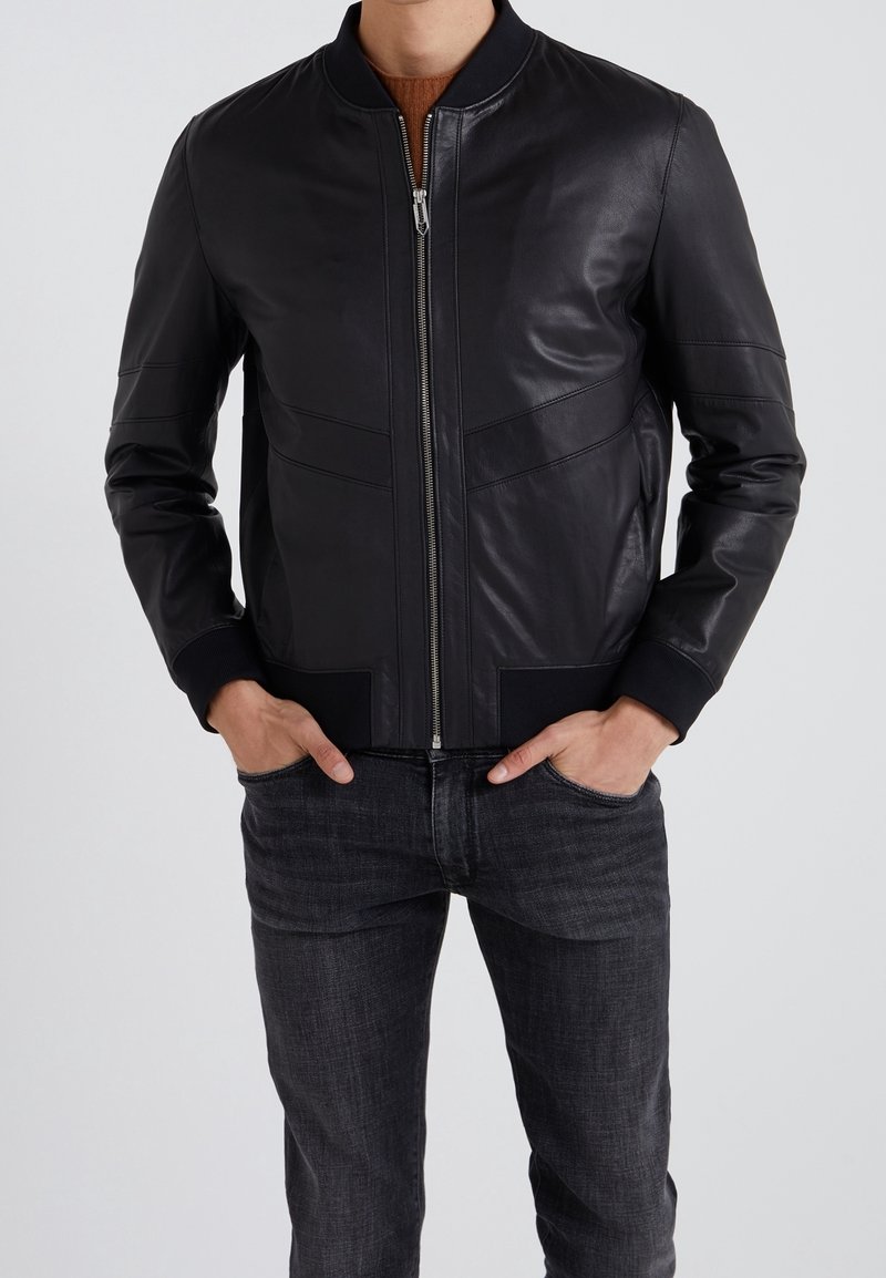 Czarna skórzana kurtka bomberka z przednim zamkiem błyskawicznym, ściągaczami przy mankietach i u dołu oraz pionowymi przeszyciami podkreślającymi strukturę. W połączeniu z ciemnymi jeansami.