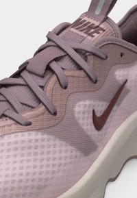 Primer plano de una zapatilla Nike malva que muestra tela texturizada, cordones y el logo Nike en color burdeos en el lateral sobre una suela blanca.
