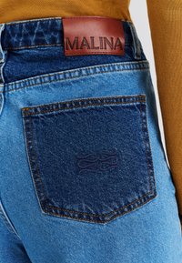 Világoskék mosású farmernadrág, sötétkék hátsó zsebbel, dombornyomott mintával, és egy barna bőr övpánt címkével, amelyen az "MALINA" felirat olvasható.