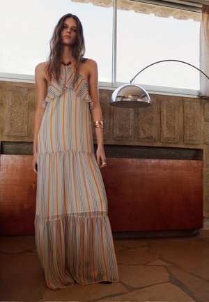 Vrouw in een mouwloze, geplooide, gestreepte maxijurk die binnen staat bij een moderne vloerlamp en een gestructureerde muur met grote ramen erboven.