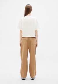 Camiseta blanca recortada de manga corta, combinada con pantalones marrones de pierna ancha que tienen dos bolsillos traseros y detalles dorados en los puños.