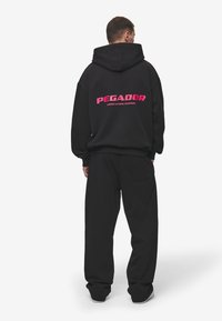 Black oversized hoodie with a pink "PEGADOR" logo on the back, paired with matching black pants. The fabric appears soft and textured.