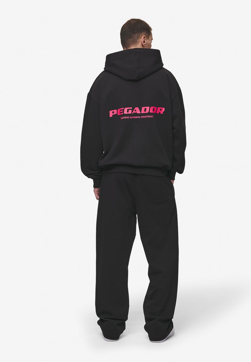 Black oversized hoodie with a pink "PEGADOR" logo on the back, paired with matching black pants. The fabric appears soft and textured.