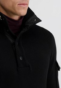 Zwarte gebreide pullover met een knoopsluiting, hoge geribbelde kraag en contrasterende gevoerde kraag. Heeft een detail aan de zijnaden.