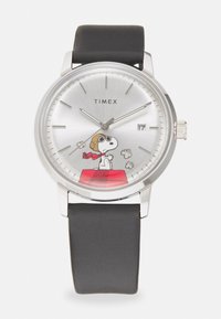 Montre Timex avec un cadran argenté, un bracelet en silicone noir et une illustration de Snoopy sur une maison de chien rouge, présentant des marqueurs d'heure clairs.