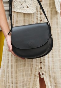 Borsa a tracolla in pelle nera con chiusura a patta tondeggiante, tracolla regolabile e cuciture visibili. Texture liscia con design minimalista.