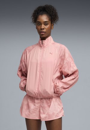 Różowa lekka kurtka z wysokim kołnierzem i elastycznymi mankietami, zestawiona z dopasowanymi krótkimi spodniami z subtelnym wzorem. Obecne logo Puma.