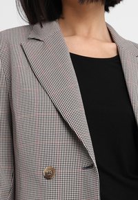 Dvouřadový blazer v černobílé kostkované tkanině s jemnými červenými akcenty. Obsahuje hnědý knoflík, klopové límce a úzký střih.