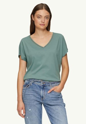 Lichtgroene T-shirt met korte mouwen en V-hals, voorzien van een gestructureerde halslijn, gecombineerd met versleten blauwe jeans. Zacht materiaal, losse pasvorm.