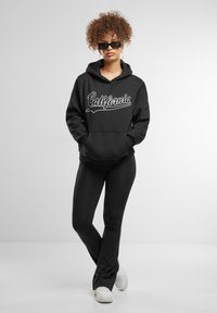 Felpa nera con grafica "California" in corsivo bianco, abbinata a leggings neri e sneakers bianche. Il modello ha i capelli ricci e occhiali da sole.