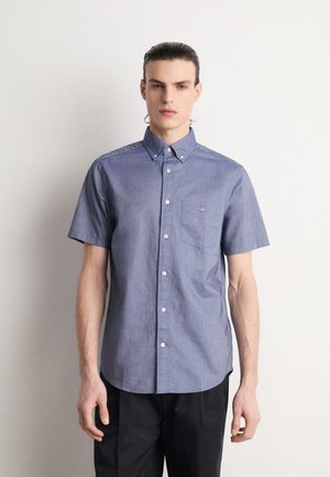Chemise en chambray bleu à manches courtes avec col boutonné, poche sur la poitrine gauche et un logo subtil. Texture lisse et coupe classique.