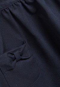 Ciemnoniebieska tkanina z małą, marszczoną kieszonką z naszywaną kokardą w lewym górnym rogu.