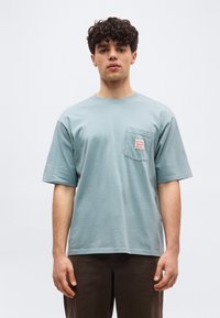 Νέος άνδρας με σγουρά μαλλιά, φορώντας φαρδιά ανοιχτό μπλε μπλούζα με μια μικρή τσέπη και σκουρόχρωμο παντελόνι, στέκεται μπροστά σε ένα απλό φόντο.