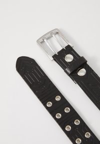 Zwarte rubberen riem met een geometrisch patroon, een zilveren metalen gesp en drukknopaccenten, voorzien van meerdere gaatjes voor aanpassing.