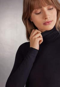 Svart polotröja med långa ärmar, figurnära design, mjuk textur och subtil lyster; bärs av en person som justerar halsringningen.