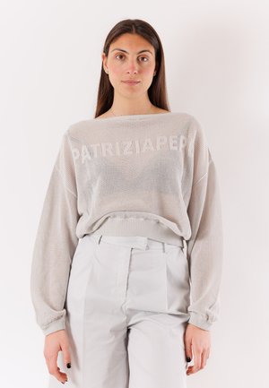 Donna con lunghi capelli scuri che indossa un maglione lavorato a maglia largo grigio chiaro con la scritta "PATRIZIA PEPE" e pantaloni grigi chiari a vita alta.