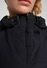 Giacca navy con un colletto alto, zip frontale, cordini regolabili e un tessuto testurizzato con dettagli di cucitura visibili.