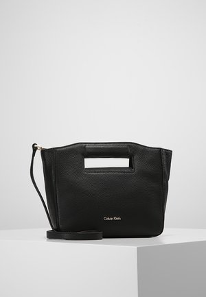 Black Calvin Klein leather handbag with cutout handle and detachable shoulder strap, placed on a white pedestal against a plain gray background.