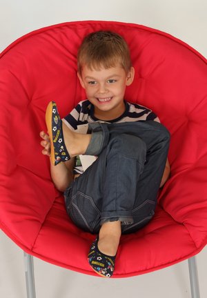 Blaue Slipper mit fußballthemed Designs, gelben Sohlen und schwarzen Akzenten. Kind sitzt in einem roten runden Stuhl und lächelt.