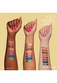Trois bras présentent des échantillons de nuances d'ombre à paupières métalliques sur des teintes de peau bronzée, moyenne et claire. Les couleurs vont des ors aux bleus, affichées en rangées étiquetées.