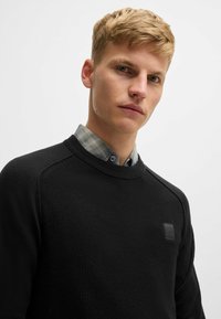 Svart stickad sweatshirt med ett strukturerat mönster, rund halsringning och raglanärmar, med en liten logotyp från varumärket på bröstet.