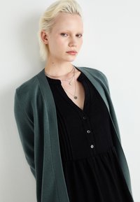 Black button-up dress with pleats, layered under a soft green cardigan. The outfit features silver layered necklaces and a smooth texture.