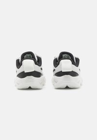 Chaussures de sport noires et blanches avec une tige en maille texturée, un talon arrondi et une semelle en caoutchouc. Présente un onglet de tirage blanc et un logo sur le talon.