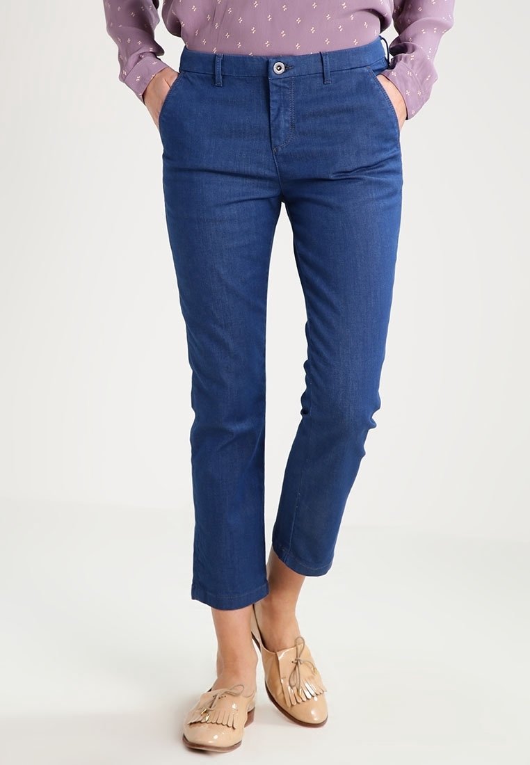 Femme portant un pantalon bleu slim raccourci, une chemise violette à manches longues et des mocassins beige à pampilles, debout les mains dans les poches.