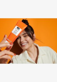 Oransje tube med Origins solkrem med en hvit bunn, med en svart logo og kremfarget etikett. Hånd som holder tuben, lys oransje bakgrunn.