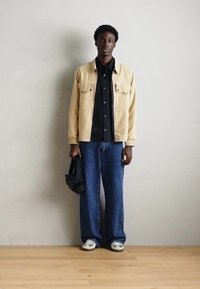Beige katoenen jack over een zwart shirt, gecombineerd met wijde blauwe spijkerbroeken en grijze sneakers. De persoon houdt een zwarte tas vast.