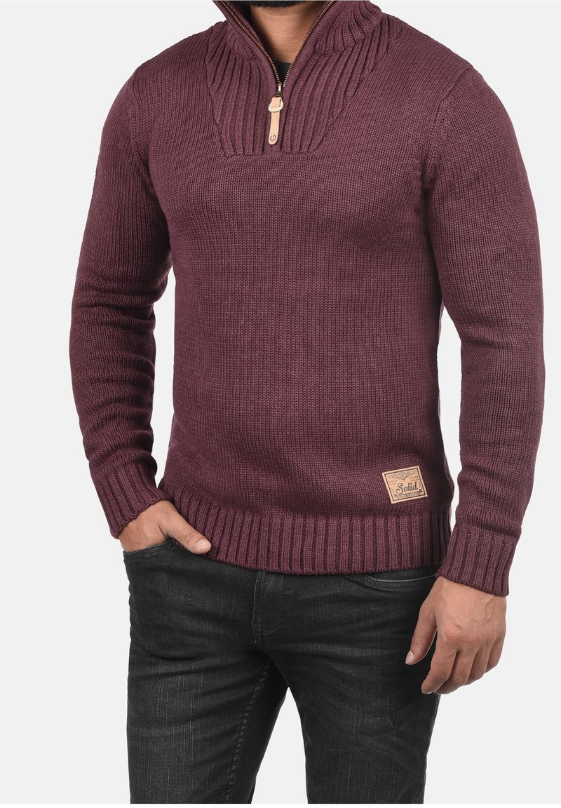 Bordeauxroter Strickpullover mit hohem Kragen und halbhohem Reißverschluss, mit gerippten Bündchen und Saum sowie einem kleinen beigen Logopatch am unteren Rand.