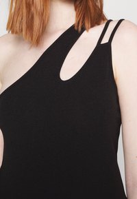 Femme aux cheveux roux mi-longs portant un haut noir asymétrique avec une découpe en forme de trou de serrure et deux fines bretelles doubles.